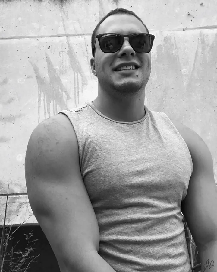 Homme en noir et blanc, portant des lunettes de soleil et un débardeur gris, souriant