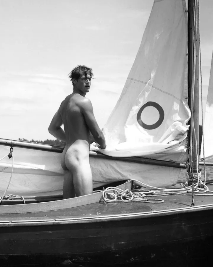 Homme en noir et blanc, torse nu, debout sur un bateau à voile