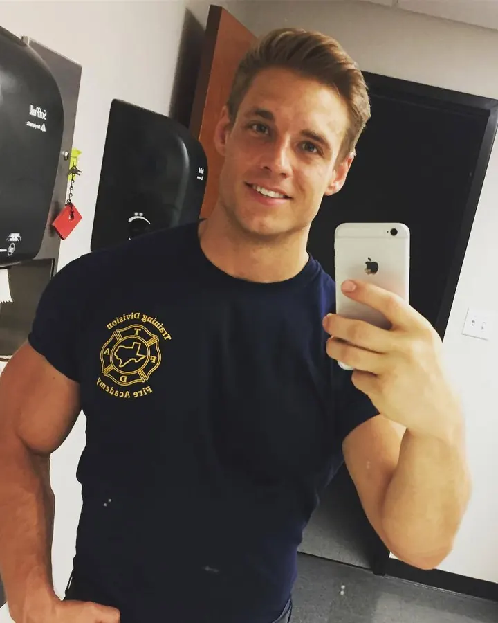 Homme souriant en t-shirt bleu marine, prenant un selfie dans un miroir, musclé, cheveux châtains clairs.