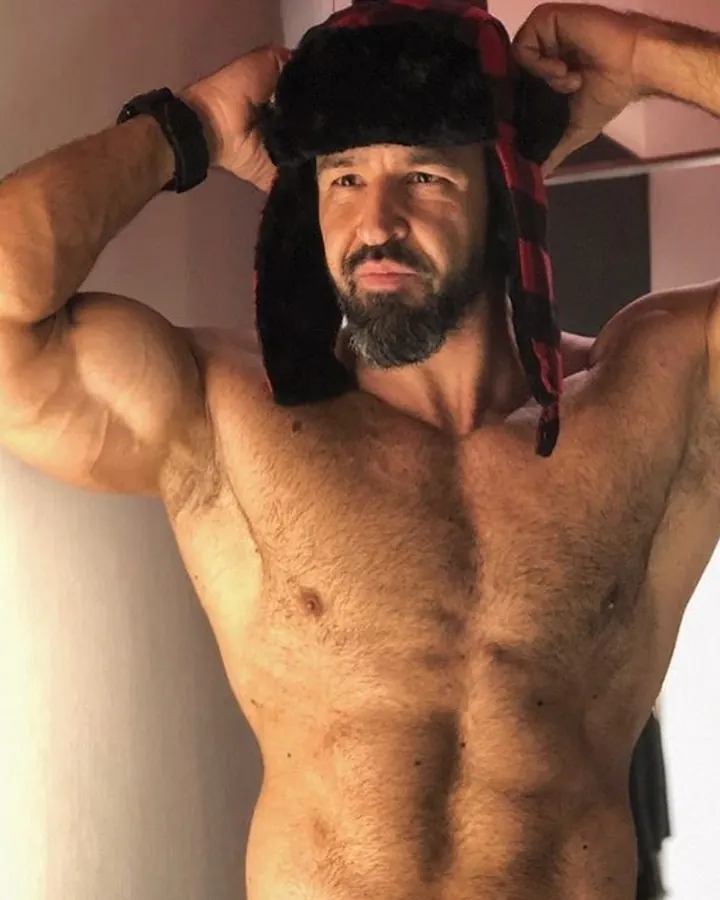Homme en Nike, jambe tendue contre le mur, chaussures noires, geste du pouce levé, chambre sombre.