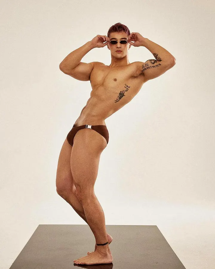 Homme torse nu, cheveux roux, tatouages sur le torse et bras, portant un string marron, posant en studio blanc, lunettes de soleil.