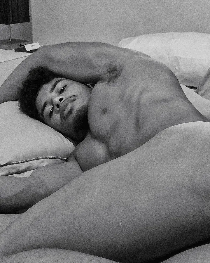 Homme torse nu, cheveux foncés, regard intense, assis sur un lit, couvert par une couette blanche, photo en noir et blanc.