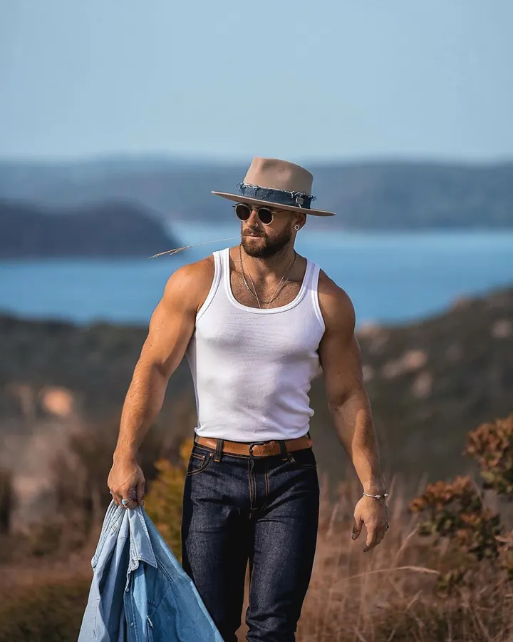 Homme barbu en débardeur blanc et jean, portant un chapeau de cowboy, marchant en extérieur près d’un lac, regard pensif.