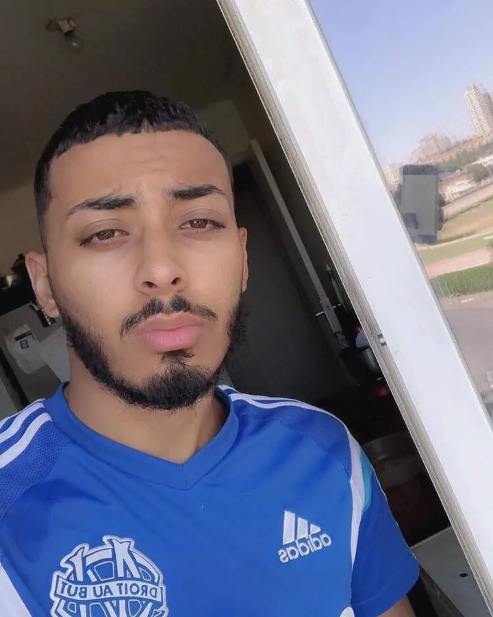 Homme torse nu dans une pièce, portant un t-shirt Adidas bleu, regardant l'appareil