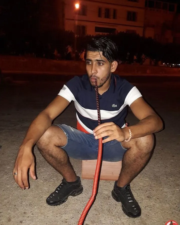 Jeune homme assis sur un bloc en bois de nuit, portant un t-shirt Lacoste et des shorts, tenant un tuyau rouge dans la bouche