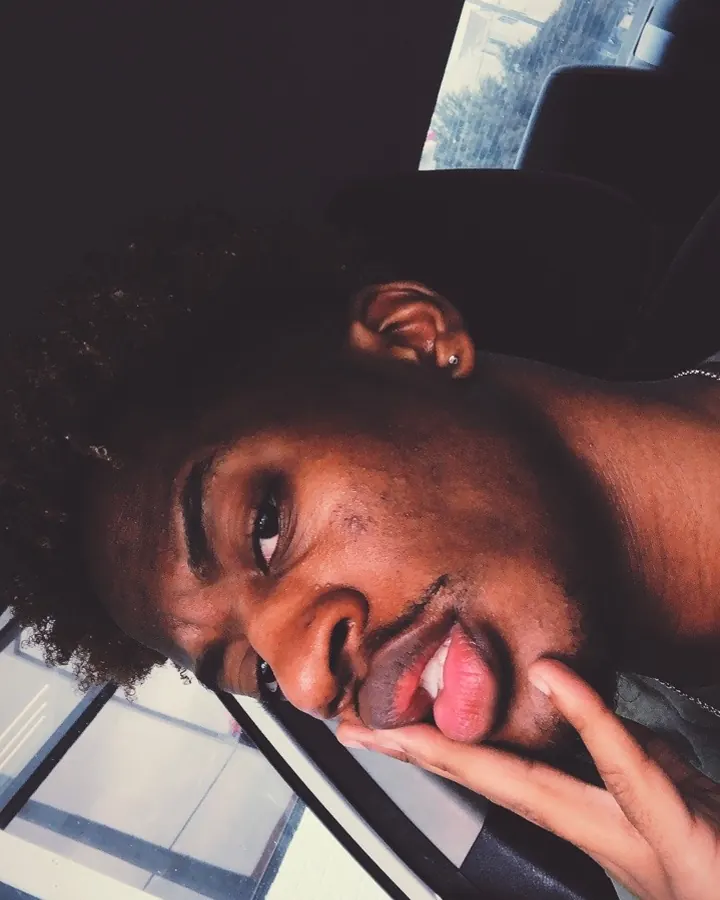 Homme noir en chemise noire, portant des lunettes de soleil, une couronne verte, un haut rayé noir et blanc, un pantalon rayé noir et blanc, les mains sur le torse, debout sur une colline herbeuse.