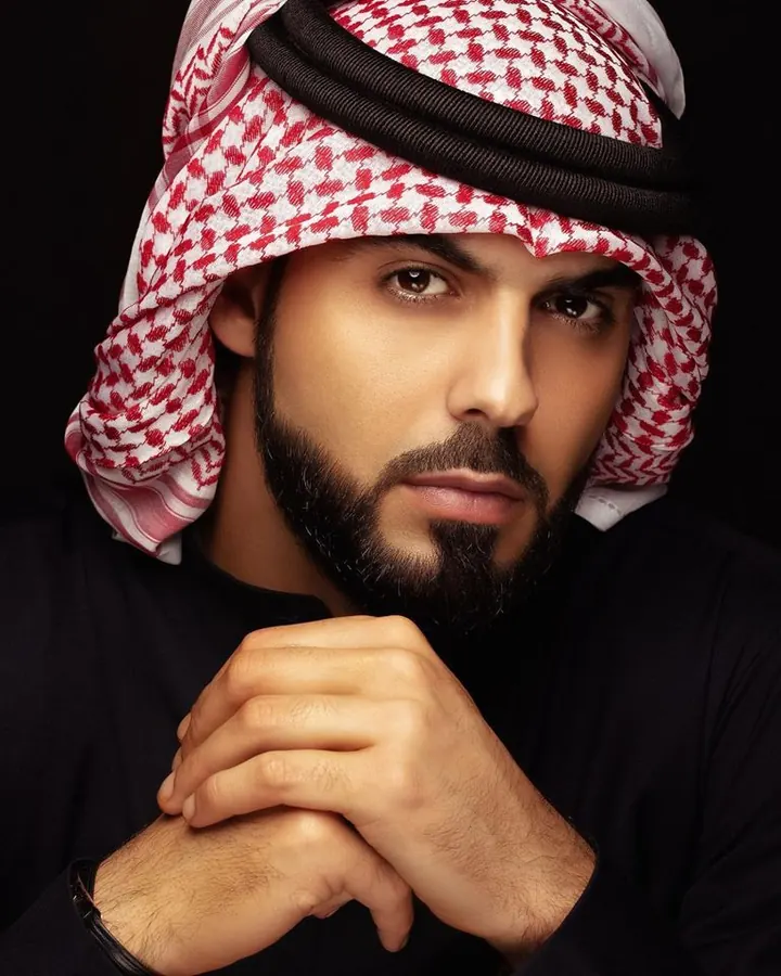 Homme en tenue traditionnelle, foulard rouge et blanc, mains jointes, regardant l'appareil photo avec intensité.