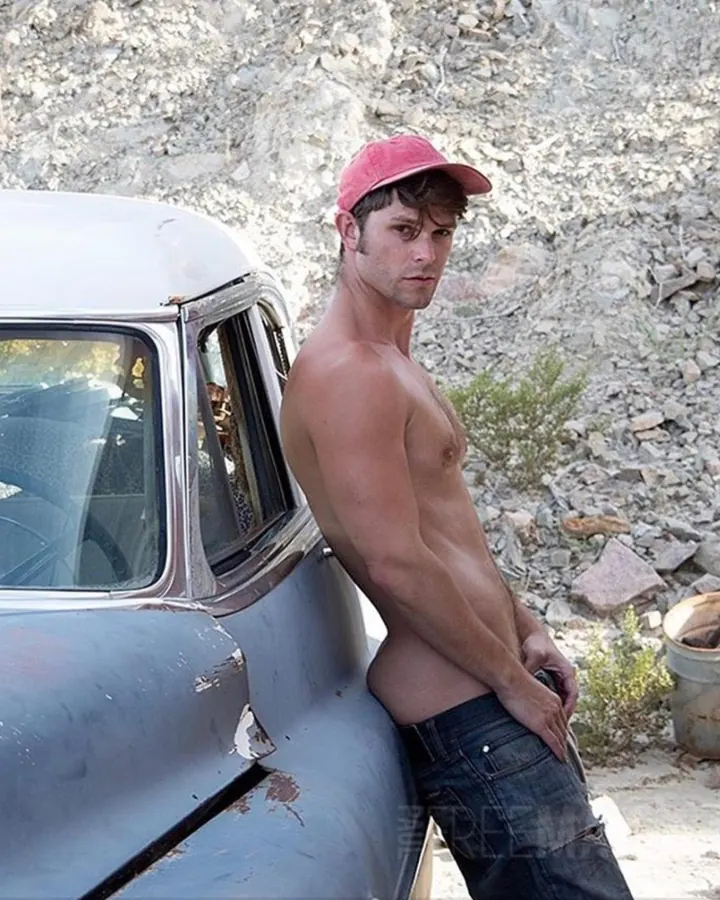 Homme torse nu portant un bonnet rose, appuyé contre une vieille voiture dans un décor rocailleux.
