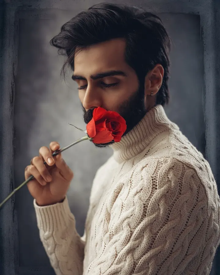 Homme aux cheveux foncés et barbe, portant un pull beige, tenant une rose rouge près de son visage.
