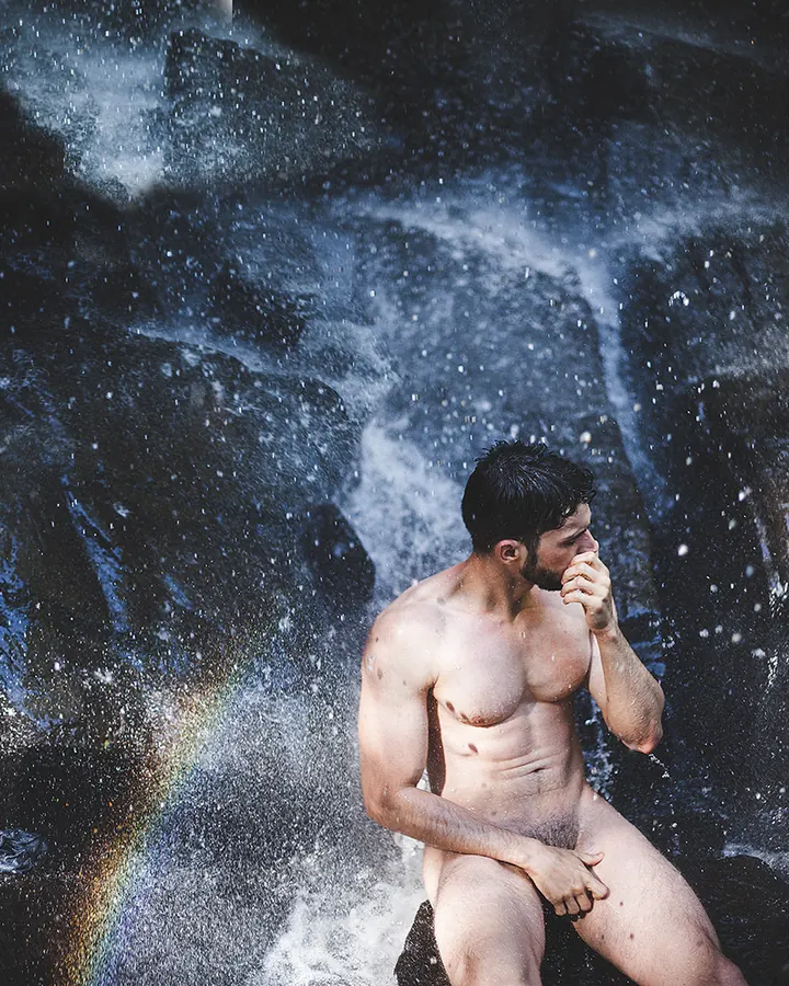 Homme torse nu, barbu, assis sous une chute d'eau, main sur le visage, arc-en-ciel visible dans le jet d'eau.