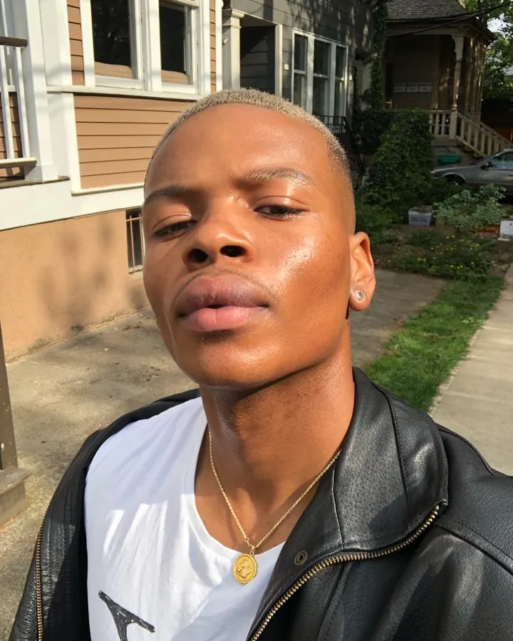 Homme africain aux cheveux courts, portant une veste en cuir, regardant vers le côté dans une rue ensoleillée.