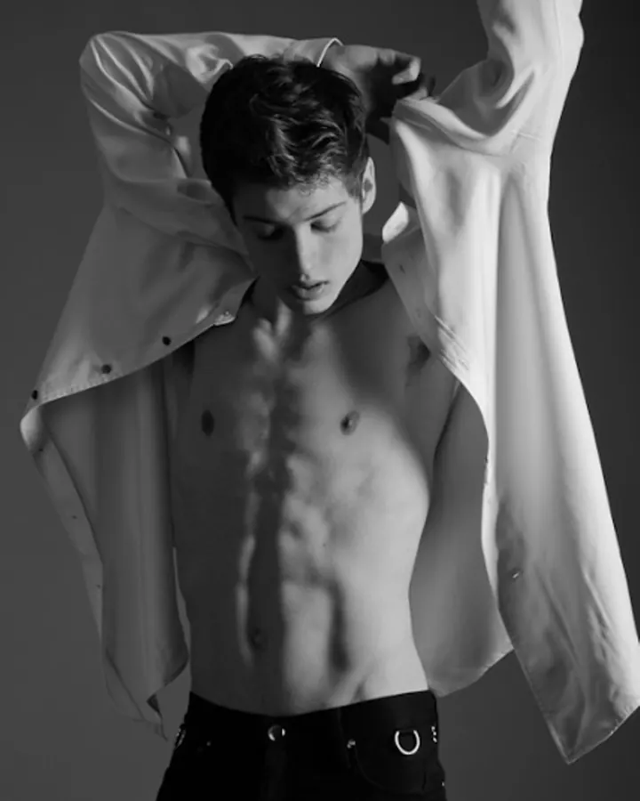 Jeune homme en chemise blanche ouverte, torse nu, regardant vers le bas, sur fond sombre en noir et blanc