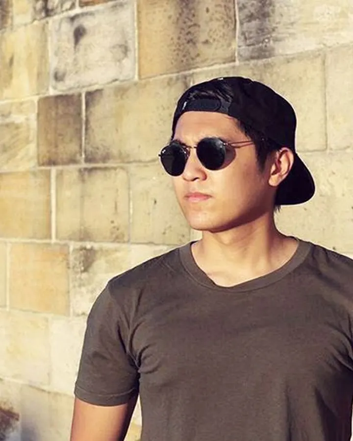 Homme blanc, en t-shirt sombre et casquette, debout contre un mur de briques.
