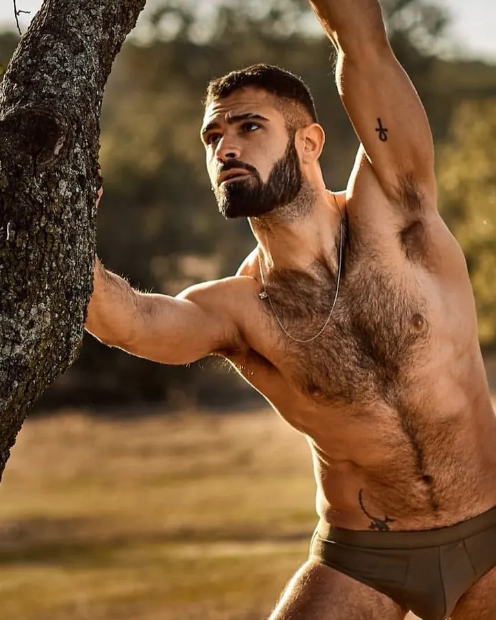 Homme blanc, barbu, torse nu, tenant un arbre dans un paysage ensoleillé.
