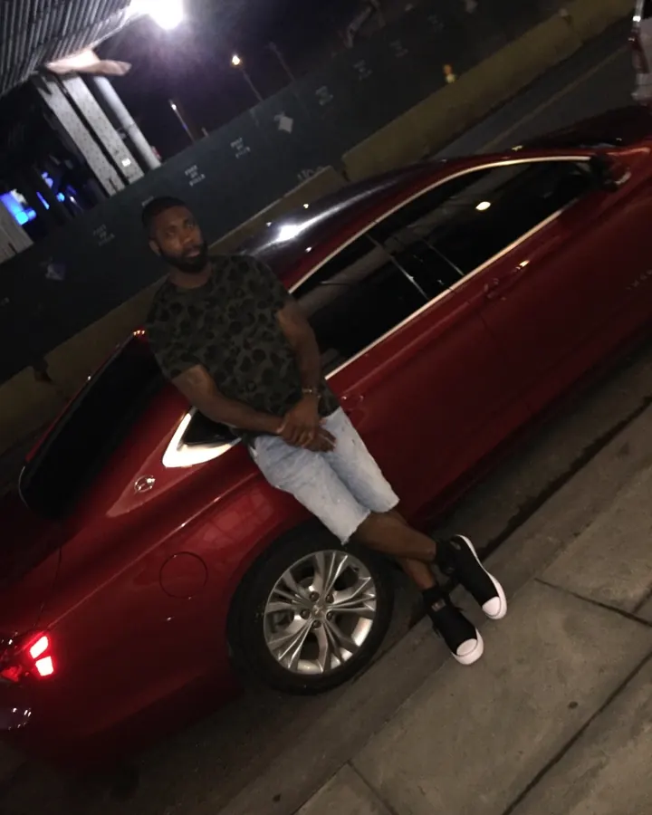 Homme noir, barbu, en t-shirt camouflage, assis sur une voiture rouge la nuit.