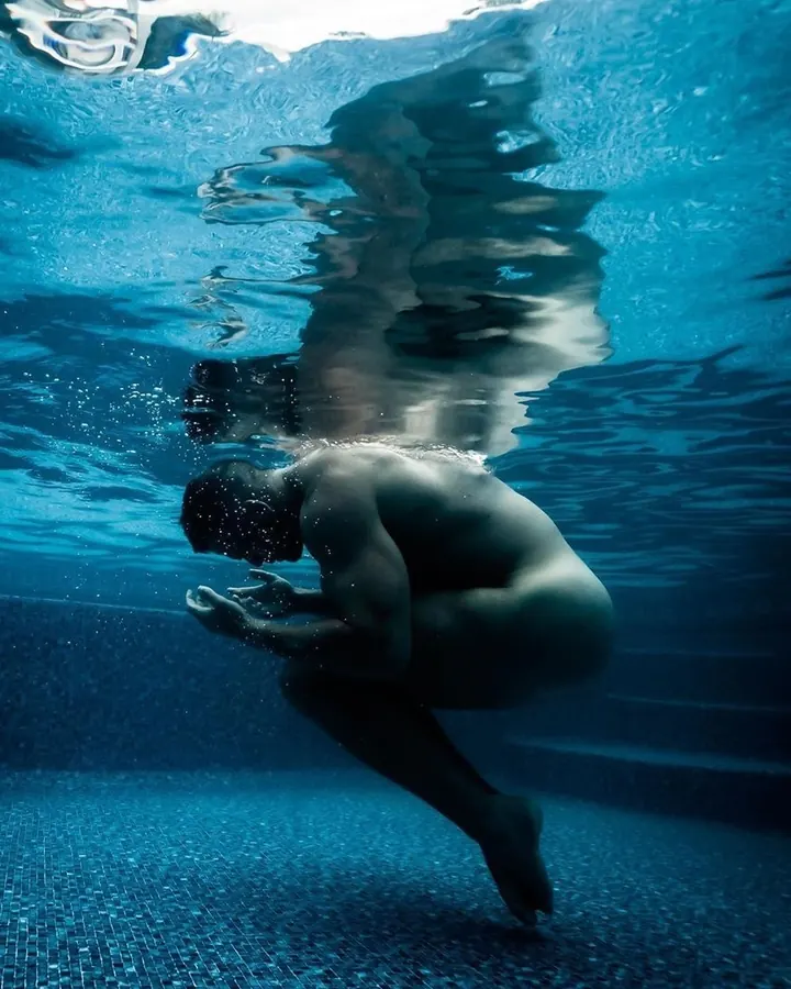 Homme torse nu sous l'eau, plongeant vers le fond d'une piscine bleue, reflets lumineux sur sa peau.