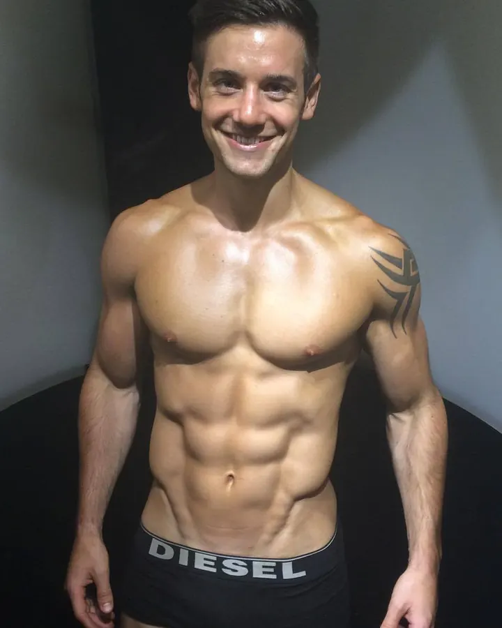 Homme torse nu, souriant, en caleçon noir Diesel, tatouage visible sur l'épaule, pose devant un fond sombre.