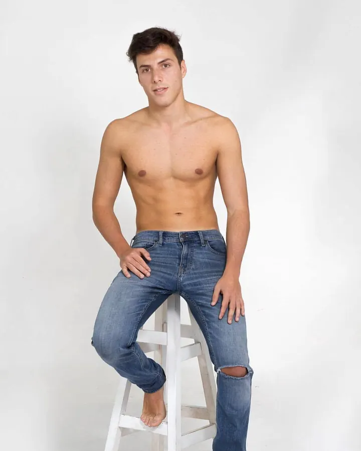 Jeune homme torse nu, assis sur un tabouret blanc, portant un jean déchiré, regardant directement l'objectif