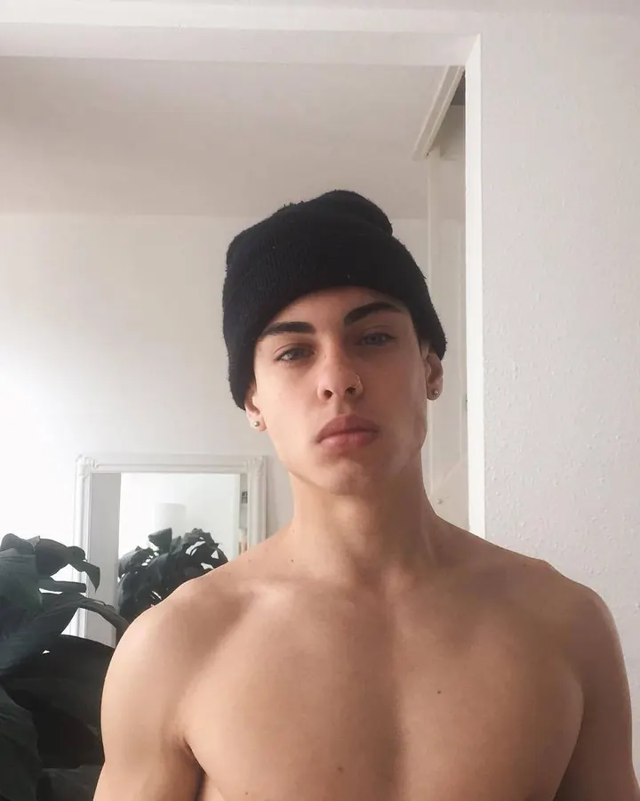 Jeune homme torse nu, cheveux bruns, regardant directement l'objectif, dans un cadre sombre