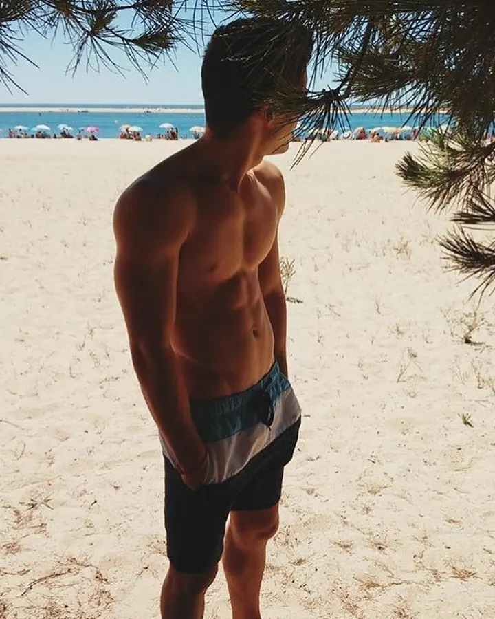 Homme torse nu, en short de plage, debout sur une plage, regardant vers la mer.
